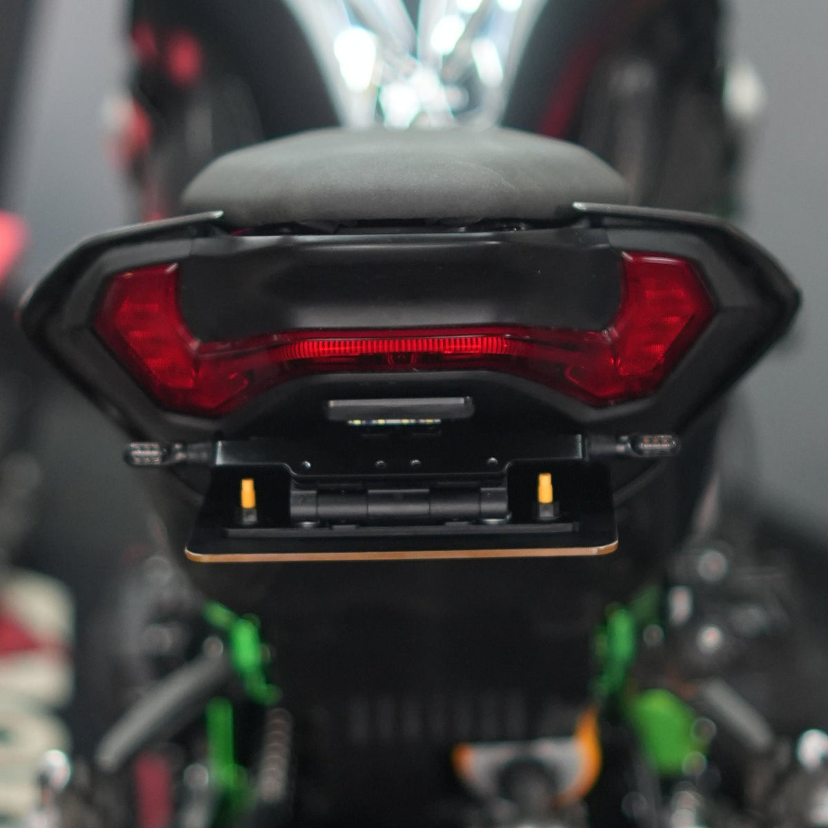 Close-up rear view of a black motorcycle with a red taillight and chrome exhaust, photographed in a workshop setting with blurred tools and equipment in the background.