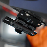 Close-up of a black metal mounting bracket or hinge component attached to a dark blue-gray panel, with a blurred orange and gray background.